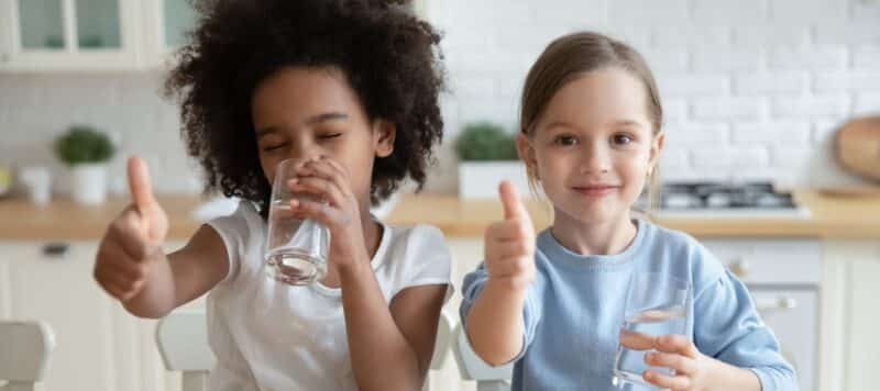 two young kids sitting at a counter drinking purified water with their thumbs up in approval