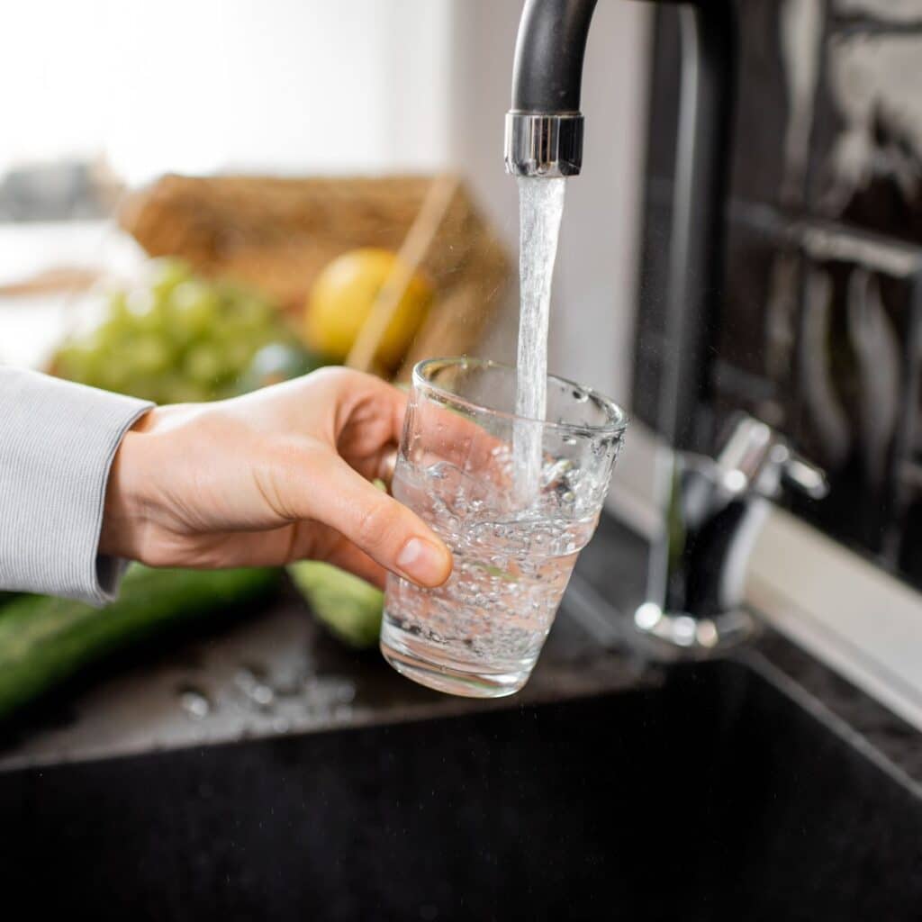 A person fills a glass with water from a kitchen faucet, with cucumbers and fruit in the background