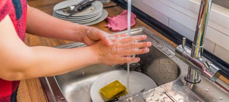 A person's soapy hands are washing in a kitchen sink filled with dirty dishes and a yellow sponge.